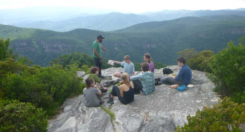 A group of people sit on a rock overlook. Below them is a vast green mountainous area. 